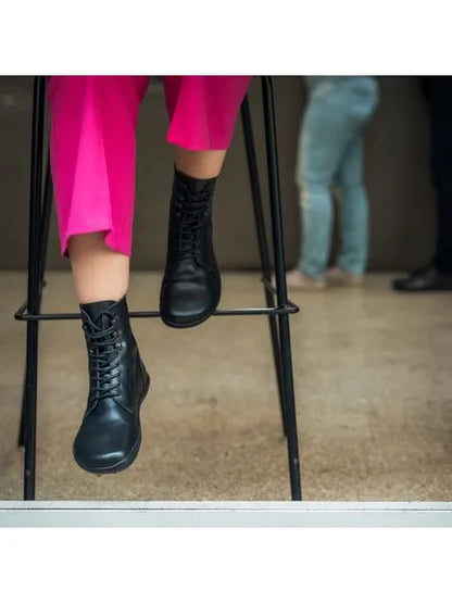 Detalhe de uma pessoa sentada num banco alto, vestindo calças cor-de-rosa vibrantes e as botas Angles Orpheus zip winter pretas.