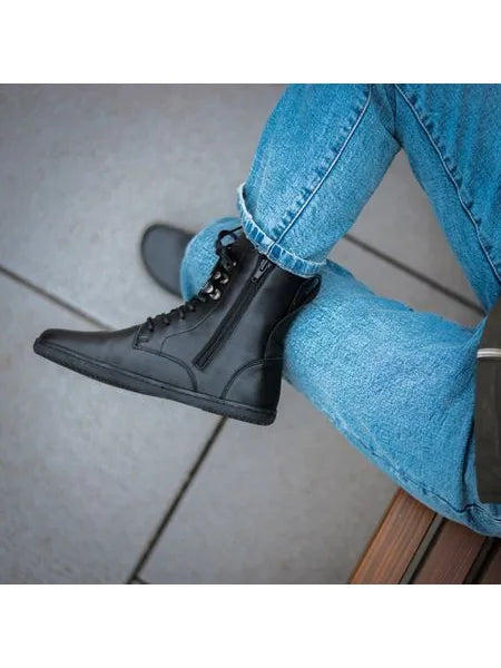 Pessoa sentada, com calças de ganga dobradas, mostrando a bota preta Angles Orpheus zip winter com fecho lateral visível.