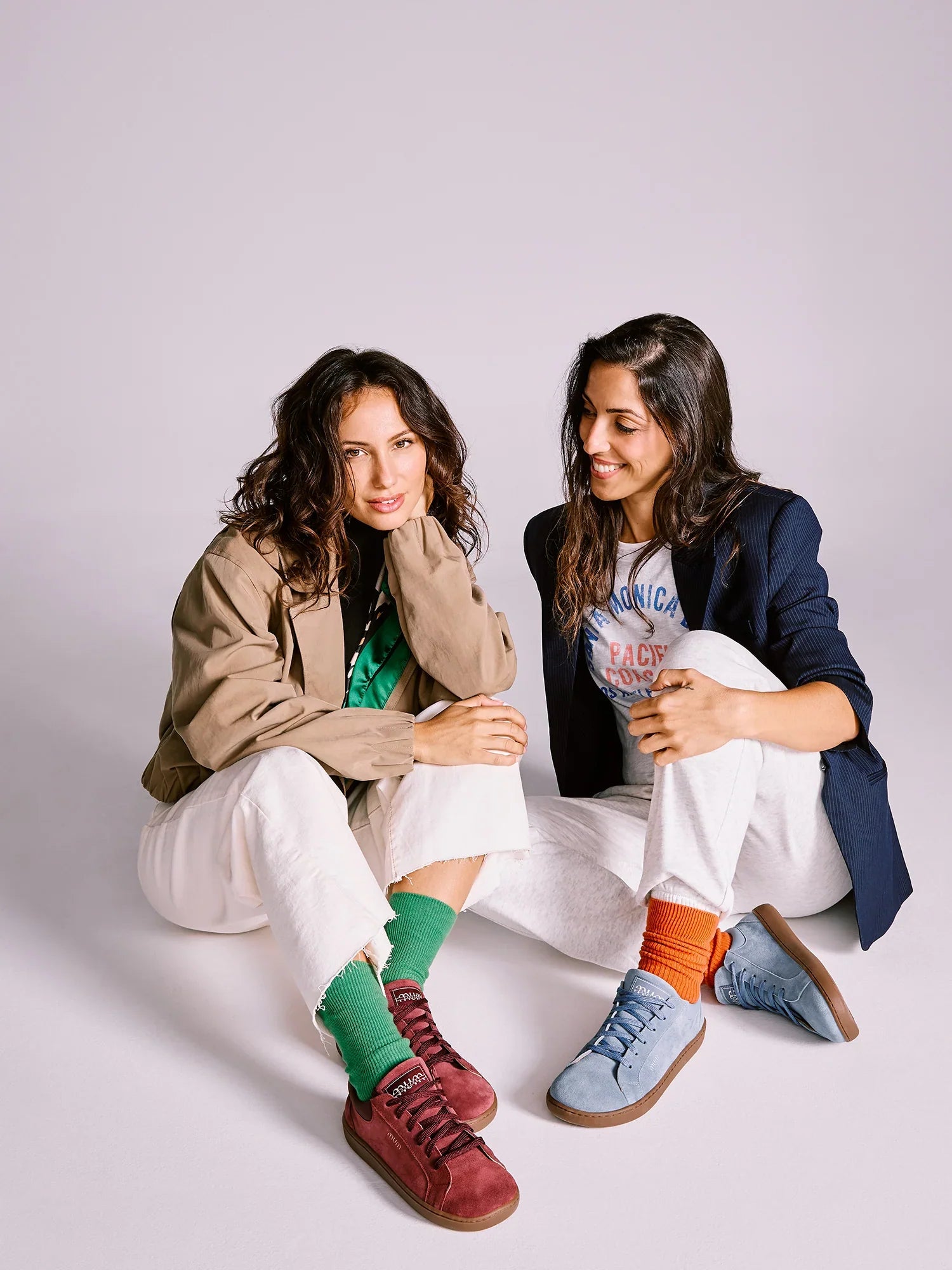 Duas mulheres sentadas, uma a usar sapatilhas MUM Barefoot Diana Wine bordô e outra sapatilhas azuis.