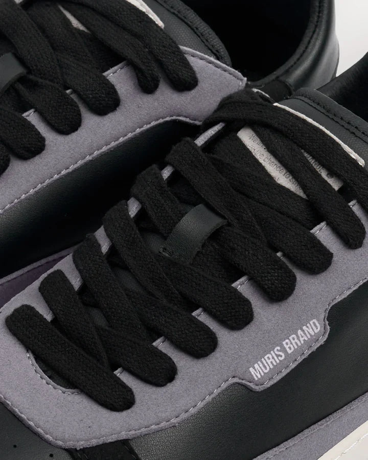 Detalhe em primeiro plano da sapatilha barefoot Muris Majorca Black, mostrando atacadores pretos, painéis cinzentos e a inscrição "MURIS BRAND" em relevo.