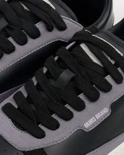Detalhe em primeiro plano da sapatilha barefoot Muris Majorca Black, mostrando atacadores pretos, painéis cinzentos e a inscrição "MURIS BRAND" em relevo.
