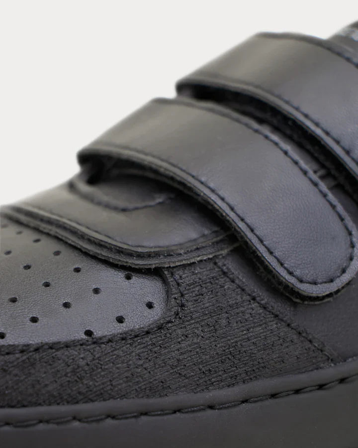 Detalhe em close-up da sapatilha Muris Petra Junior Black, mostrando o fecho de velcro duplo e a textura dos materiais sustentáveis na zona da biqueira.