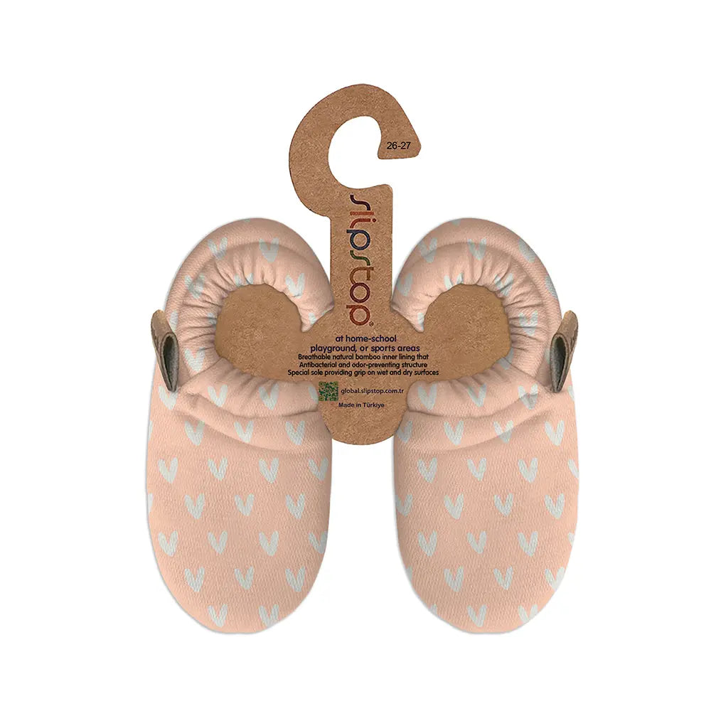 Par de pantufas Slip Stop Booties cor de rosa claro com padrão de corações brancos, suspensas em embalagem de cartão com logótipo e informações do produto.