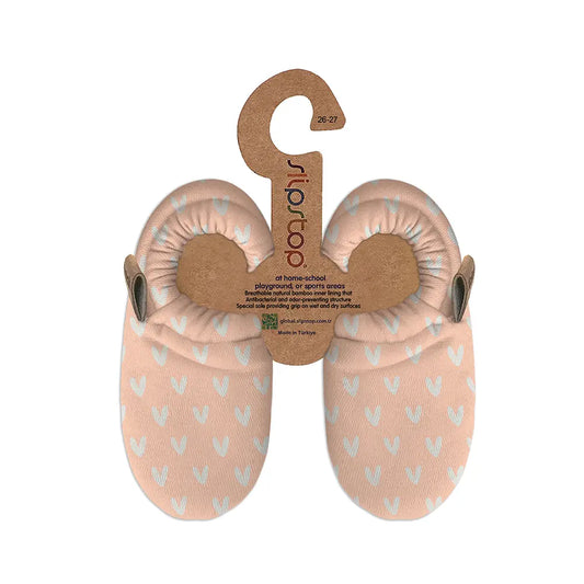 Par de pantufas Slip Stop Booties cor de rosa claro com padrão de corações brancos, suspensas em embalagem de cartão com logótipo e informações do produto.